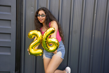 Obraz na płótnie Canvas Cheerful beautiful young indian woman wearing casual cloths showing golden balloon number twenty six.