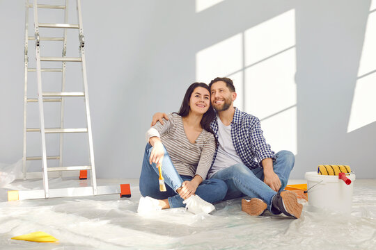 Smiling happy couple painting the wall of their new home holding paint brush near ladder. Married man and woman sitting on the floor and planning repair renovation preparing to move into a new flat.