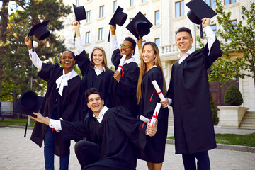 Young multinational people university students dressed in black gown raise their hats rejoicing in successful completion higher education with excellent grades standing in front of college building
