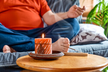 Overweight man practicing yoga and meditation at home sitting relaxed in lotus pose on the couch in front of aromatherapy burning candle. Mindful meditation concept. Wellbeing.