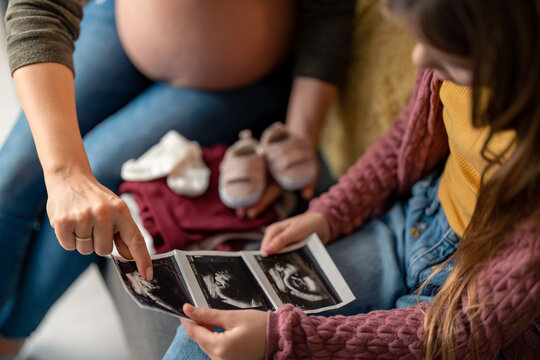 Cropped shot of unrecognizable pregnant woman sitting next to her daughter pointing on ultrasound picture, explaining to her how the baby looks right now, holding pair of non focused pale pink shoes.