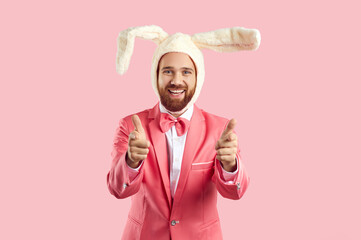 Funny guy in rabbit costume pointing his fingers at you. Happy joyful bearded young man wearing fluffy toy Easter bunny ears standing against pink studio background, smiling and pointing at camera