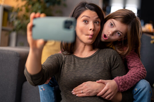 Mother and daughter making funny faces, grimacing, enjoying their time together while taking selfies at home.