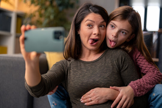 Playful mother and daughter making funny faces, grimacing, sticking out tongue, enjoying their time together while taking selfies at home.