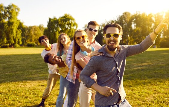 Funny and cheerful joyful company of young friends have fun together on green lawn in park. Multiracial people have fun and dance together walking in row and holding each other's shoulders together.