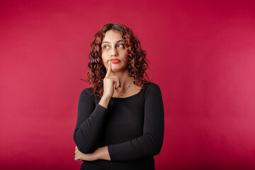 Fototapeta premium Young beautiful woman wearing black ribbed dress isolated over red background undecided, purses lips and touches with index finger. Looks at the empty copy space and tries to decide.