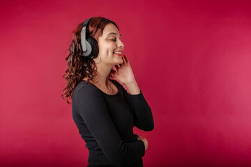 Happy woman wearing black dress standing isolated over red background eyes closed, listens to music by touching headphone and feels the music. Enjoys and laughs a lot while listening to favorite music