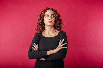 Portrait of young redhead woman wearing black dress standing isolated over red background with folded arms looks at the empty copy space with an expression of disapproval.