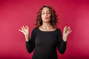 Woman standing isolated over red background keeping eyes closed, exhales, holding fingers in mudra...