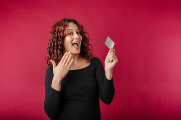 Happy redhead woman wearing black ribbed dress standing isolated over red background holding credit card, showing membership card mockup shopping payment money financial online.