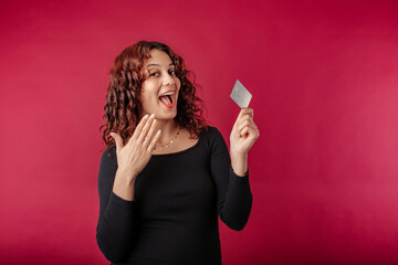 Happy redhead woman wearing black ribbed dress standing isolated over red background holding credit...