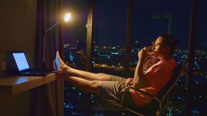 Fototapeta na wymiar Man in an apartment against background of night city. Handsome man in room luxury hotel works at laptop. Nice guy lives or freelancing works in hotel. Man sits on an office chair against large window