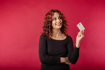 Happy redhead woman wearing black dress standing isolated over red background holding a credit card looks up with a thoughtful expression. Trying to decide what to buy. Credit card advertising concept