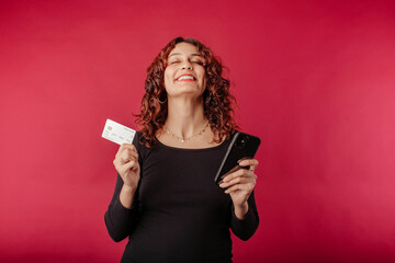 Naklejka premium Happy redhead woman standing isolated over red background holding a credit card and a cell phone poses with a very happy smile. Experiencing the happiness of shopping. Eyes are closed.
