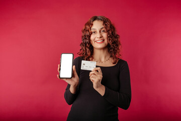 Portrait of redhead woman standing isolated over red background holds credit card and phone with blank white mockup screen side by side and shows it to the camera. Looks at the camera with a smile.