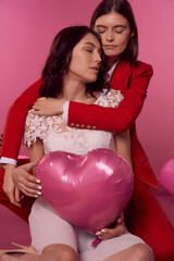 Fototapeta na wymiar Valentine's day concept. Studio portrait of a lgbtq couple holding pink a heart balloon and sitting on the floor on a pink background. One girl is in a white dress, the second woman is in a red suit.