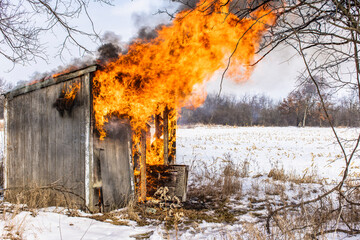 A two door outhouse or small shed on fire in the winter with large flames. - Powered by Adobe