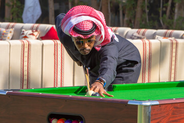 The Saudi Man wearing the traditional dress "SHEMAGH scarf" red and white color with black dress he is playing Pool game in the garden with green grass