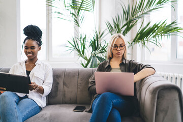 Diverse women using gadgets while sitting on sofa near window