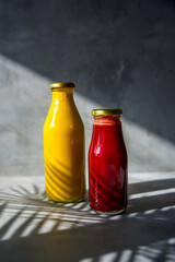 Two bottles of healthy raw cold pressed juice. One bottle is red juice and one bottle is yellow juice. There is a palm leaf shadow across the bottles.