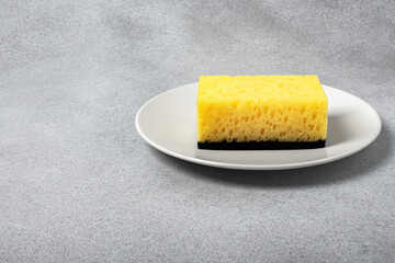Yellow sponge for dishwashing on a white plate on a neutral gray background. Gentle dishwashing. House cleaning.