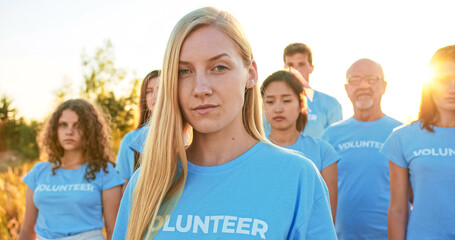 Obraz premium Young attractive woman with a group of eco activists in the background. Pretty Caucasian female volunteer looking at camera and saying: I Am a Volunteer. Volunteering, environmental conservation.