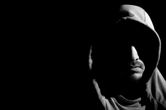 Black and white horizontal portrait shot of mysterious young bearded hooded man with his face covered in shadows isolated on dark black background with copy space. Concept of anonymous person.