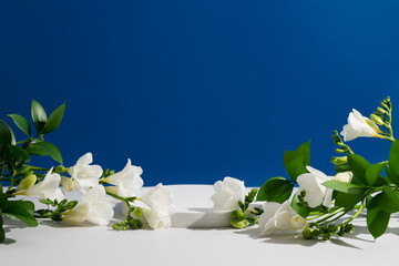 Empty white podium with freesia flowers and green leaves on dark blue background. Showcase for...