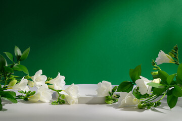Pedestal for product presentation. Empty white podium with freesia flowers and naturals leaves and deep shadows on green background. Showcase for beauty cosmetic advertising. Still life. Front view. - obrazy, fototapety, plakaty