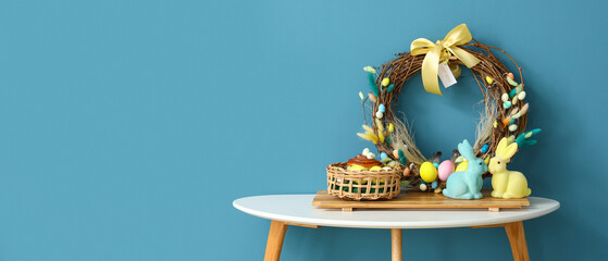 Basket with Easter eggs, cake, beautiful wreath and bunnies on table near blue wall. Banner for design