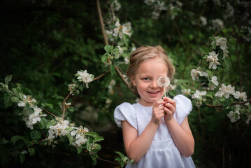 A beautiful girl in a white dress covers her eye with a dandelion flower