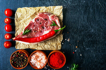 One ribeye steak raw meat barbecue beef, hot peppers, spices, tomatoes, salt, paprika, pepper mix, rosemary on paper on a black table