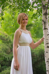 Naklejka na ściany i meble blonde beautiful natural girl with blonde hair in a white dress in nature, on the street, in the garden by a tree, with makeup and hairstyle, greek woman in a white ethnic historical dress, retro