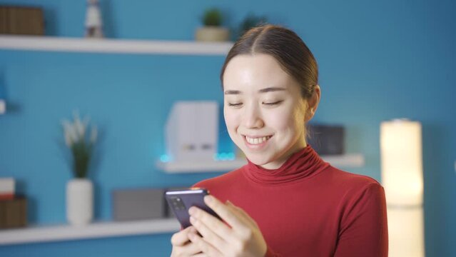 Asian woman looking at phone happily at home.
Young asian woman alone at home surfing internet on phone, talking with friends, looking at news and happy, in a good mood.
