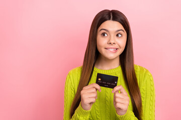 Portrait of cute positive girl hold debit card biting lip look empty space isolated on pink color...