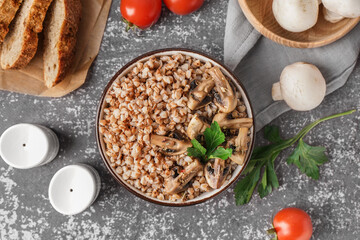 Bowl of tasty buckwheat porridge with mushrooms, parsley and fresh tomato on grey table