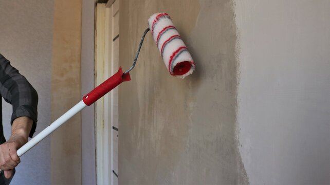 red and white roller on a long handle in the process of applying a layer of glue on a plastered wall in a home room, priming room walls with a homemade long roller and copy space for pasting