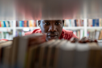 African American man standing between bookshelves in university library, picking up book from...