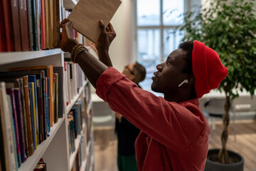 Concentrated young African American guy picking book for preparing for exam, project in literature or test from bookshelves in university campus. Man college student in library. Education and studying