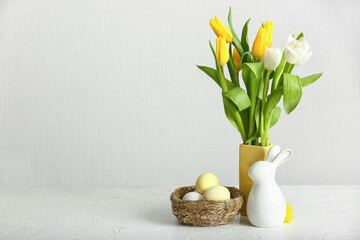 Vase with beautiful tulip flowers, nest of Easter eggs and ceramic bunny on light background
