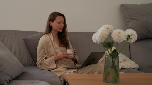 Side view of young woman in bathrobe is sitting on sofa drinking coffee and making video call using a laptop in the foreground of bouquet of fresh flowers
