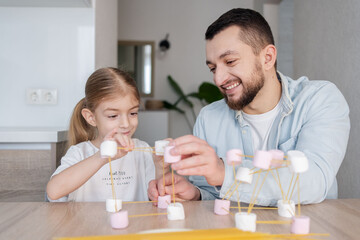 Cute child girl with father playing and creating with marshmallows and spaghetti . Child...