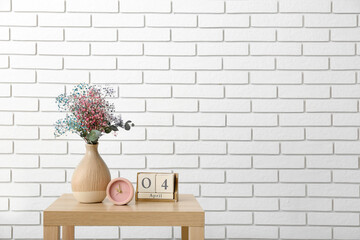 Vase with beautiful gypsophila flowers, cube calendar and alarm clock on wooden table near light brick wall