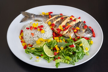 Baked dorado fish with rice garnished with salad and vegetables on a white plate, close-up.