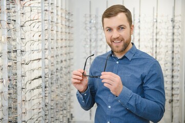 In Optics Shop. Portrait of male client holding and wearing different spectacles, choosing and trying on new glasses at optical store. Man picking frame for vision correction, closeup.
