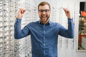 In Optics Shop. Portrait of male client holding and wearing different spectacles, choosing and trying on new glasses at optical store. Man picking frame for vision correction, closeup.