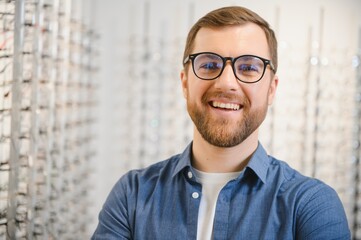 Satisfied Customer. View of happy young male client wearing new glasses, standing near rack and showcase with eyewear. Smiling man trying on spectacles
