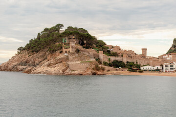 Fototapeta na wymiar Panorámica de la playa de Tossa de Mar en Girona con el castillo sobre la montaña en el fondo bajo un cielo especial con nubes.