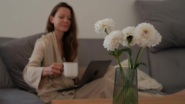 Blurred woman in a bathrobe is drinking coffee and using a laptop, focus on a fresh flowers in a vase on foreground