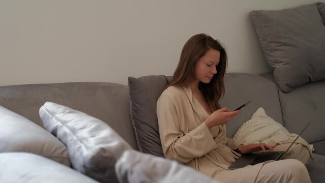 Young woman in bathrobe recording voice message using smartphone while sitting on sofa at home with laptop